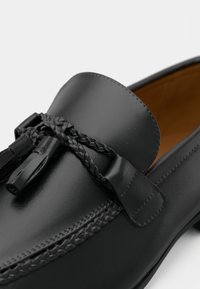 Fekete bőr loaferek sima felülettel, fonott díszítéssel és lapos talppal. Nagy pánttal és dekoratív fémrészekkel rendelkezik.