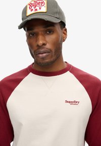 Beanie met een rechthoekig logo met "Super D.R.Y." in het rood op een beige achtergrond, gecombineerd met een crèmekleurige en bordeauxrode raglan sweatshirt.