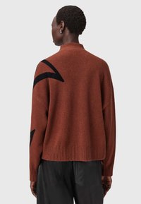 Sweter w kolorze rdzy wykonany z miękkiego materiału, z wysokim kołnierzem, ribbowanym dołem i czarnym abstrakcyjnym wzorem na lewym ramieniu.