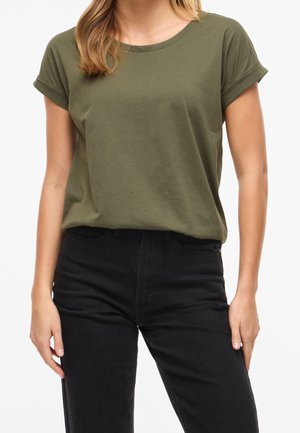 Femme portant un t-shirt vert olive à manches courtes rentré dans un jean noir taille haute, debout devant un fond blanc.
