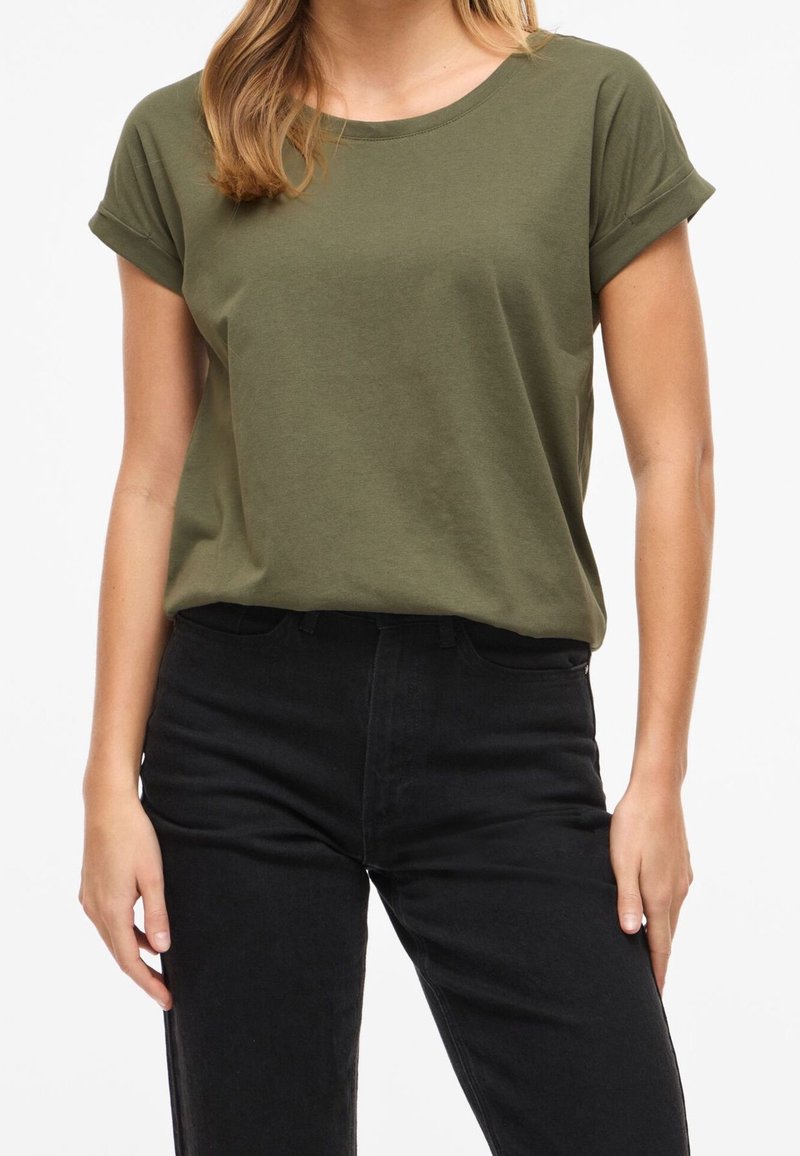 Femme portant un t-shirt vert olive à manches courtes rentré dans un jean noir taille haute, debout devant un fond blanc.