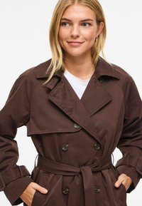 Manteau trench marron à double boutonnage, grand col et taille ceinturée ; détails de boutons et texture de tissu lisse.