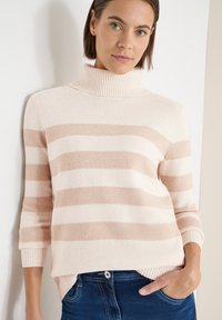 Pull crème avec larges rayures beige, doté d'un col roulé côtelé et de manches trois-quarts, associé à un jean bleu foncé.