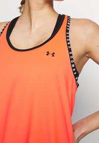 Femme portant un débardeur orange Under Armour avec un liseré noir et des bretelles de marque, main reposant sur la hanche, sur un fond uni.
