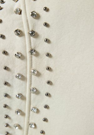 Beige stof met een verticale rij metalen studs en heldere rhinestones, wat textuur en accent aan het kledingstuk toevoegt.