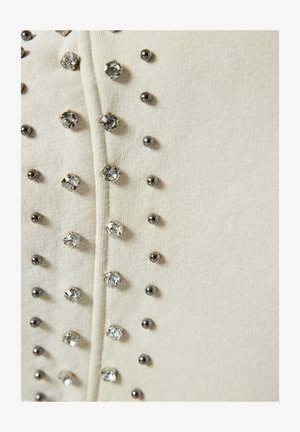 Bēšs audums ar vertikālu rindu metāliskiem kniedēm un caurspīdīgiem rhinestones, pievienojot tekstūru un akcentu apģērbam.