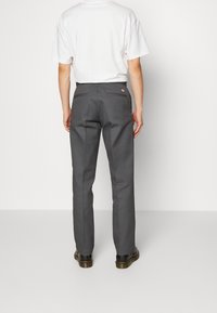 Donkere grijze broek van een gestructureerde stof. Beschikt over achterzakken en een klein logo-label aan de rechterachterkant. Draagt een wit T-shirt.