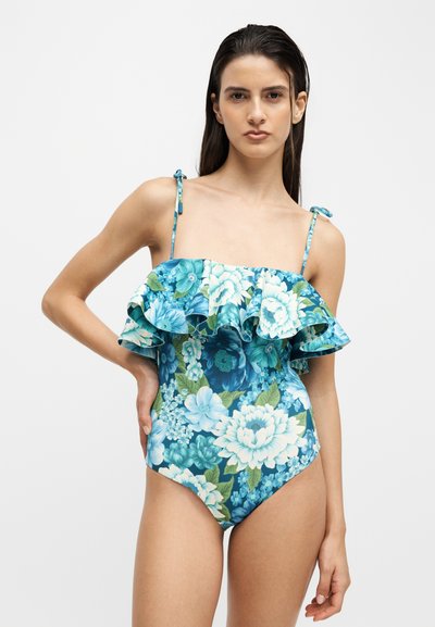 Jeune femme aux longs cheveux foncés porte un maillot de bain une pièce bleu à fleurs avec un col volanté et de fines bretelles sur fond blanc.