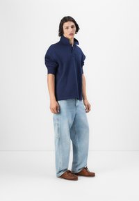 Suéter azul marino con cuello alto y tapeta de tres botones, combinado con pantalones vaqueros sueltos de color azul claro y zapatos mocasín marrones.