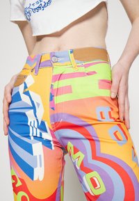 Personne portant un pantalon taille haute avec des motifs géométriques abstraits colorés et un crop top blanc partiel, mains posées sur les hanches, torse visible.