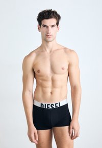Schwarze Boxershorts mit einem weißen "DIESEL"-Bund, aus glattem Material, enganliegendem Design und mittelhohem Schnitt, getragen von einem männlichen Modell.
