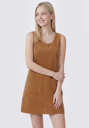 Jonge vrouw met lang blond haar die een mouwloze bruine corduroy jurk met twee voorzakken draagt, glimlachend en naar de zijkant kijkt.