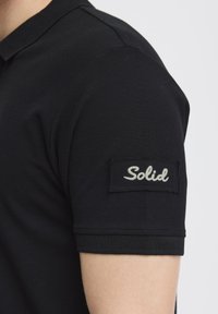 Černé polo tričko s texturovanou tkaninou a přiléhavým límečkem. Obsahuje vyšitou nášivku "Solid" na horní části rukávu.