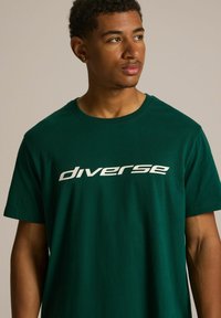 Zielona bawełniana koszulka z dużym białym nadrukiem tekstowym, który brzmi "różnorodny". Projekt zawiera klasyczny okrągły dekolt i krótkie rękawy.