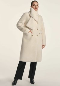 Kremowy wełniany płaszcz o miękkiej fakturze, zapinany na podwójne guziki, z złotymi guzikami oraz pasującym szalikiem, w połączeniu z czarnymi spodniami i szpiczastymi botkami.