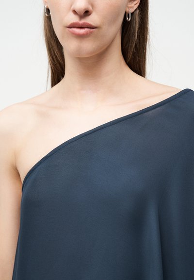 Nő sötétkék egyvállas toppban, ezüst karika fülbevalókkal, látható kulcscsonttal és semleges ajakszínnel.