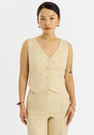 Femme aux cheveux foncés portant un gilet beige boutonné et un pantalon assorti, des boucles d'oreilles créoles dorées et des bracelets, debout devant un fond blanc uni.