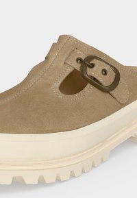 Μπεζ σουέντ παπούτσι τύπου slip-on με διακοσμητικές εγκοπές και σκούρο καφέ αγκράφα στην επάνω πλευρά, που διαθέτει χοντρή λευκή ραβδωτή καουτσούκ σόλα.