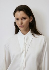 Witte button-up shirt met een grote kraag en strikdetail bij de nek. Gemaakt van een lichte stof met lange mouwen en een gladde textuur.