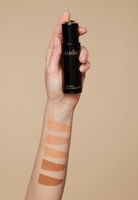 Babor Firming Serum Foundation in einer schwarzen Flasche, gehalten in der Hand. Sechs Farbproben der Foundation auf dem Arm zeigen verschiedene Beigetöne von hell bis dunkel.
