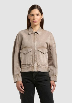 Femme portant une veste beige à fermeture éclair avec de grandes poches avant et un pantalon noir, debout devant un fond gris clair uni.