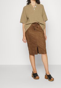 Top oversized en tricot beige avec trois boutons, associé à une jupe midi en daim marron avec une fente à l'avant, portée avec des sandales à plateforme noires.