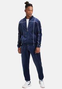 Marinblå velourträningsdräkt med vitt pipingsdetalj. Jackan har hög krage och dragkedjestängning. Byxor med elastiska muddar och dragsko i midjan.