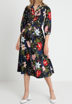 Frau trägt ein marineblaues Midikleid mit floralem Muster in Pink, Rot und Weiß, dreiviertellangen Puffärmeln und weißen Blockabsatzschuhen.