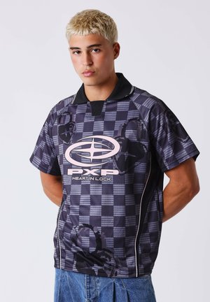 Jeune homme aux cheveux blonds courts portant un polo à carreaux foncé avec le logo "PXP Heart in Lock" et un jean bleu, debout devant un fond uni.