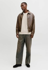 Moški stoji, oblečen v rjavo usnjeno jakno, bež srajco, izprane široke kavbojke in črne čevlje na enostavni svetli podlagi.