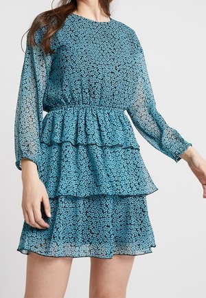 Vrouw die een blauwe jurk met lange mouwen draagt, voorzien van gelaagde ruches en een klein bloemenpatroon op een witte achtergrond.