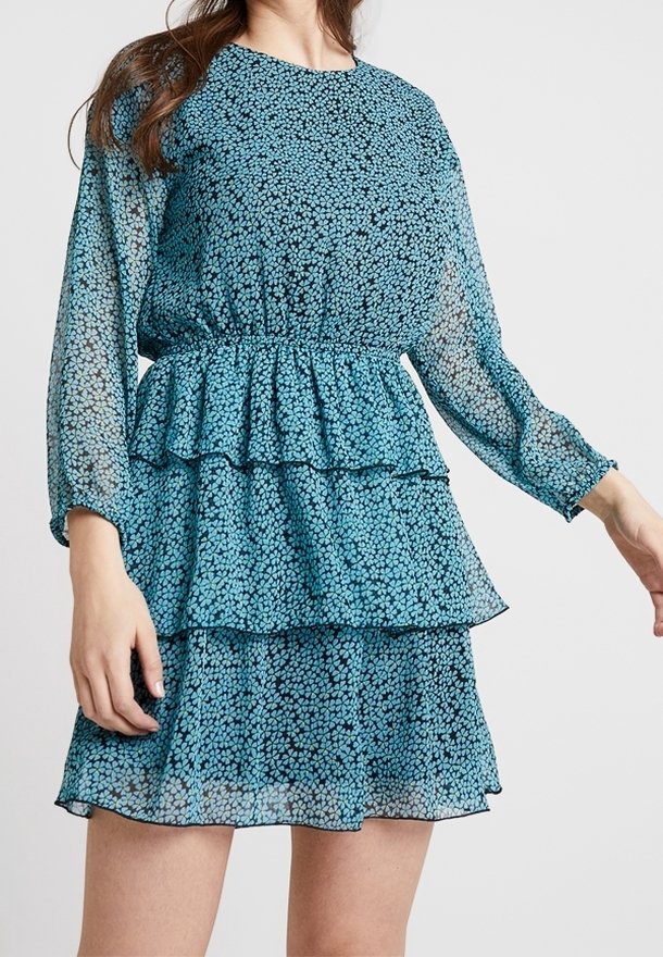 Femme portant une robe à manches longues bleue avec des volants superposés et un petit motif floral sur fond blanc.