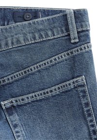 Tmavě modré džíny z denimu se přiléhavým pasem, jedním zapínáním na knoflík a viditelnými švy. Mají zadní kapsy a hladkou texturu.