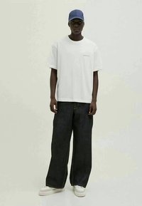 T-shirt blanche surdimensionnée, jeans larges noirs, baskets blanches et casquette bleue, se tenant contre un fond clair.