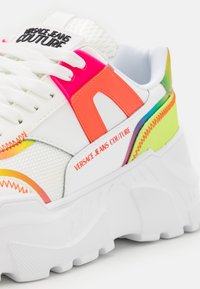 White sneaker with a chunky sole featuring neon pink and green accents, mesh texture, and "VERSACE JEANS COUTURE" branding.