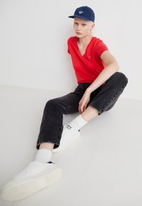 Rød V-hals t-skjorte, sorte denimjeans rullet ved ankelen, hvite joggesko, og blå baseballcap med logo. Hvite teksturerte sokker synlige.