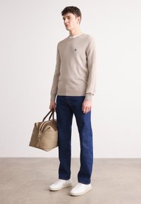 Suéter de punto beige con dobladillo acanalado, jeans azul oscuro y zapatillas blancas. Bolsa beige con detalles en marrón, sostenida en la mano derecha.