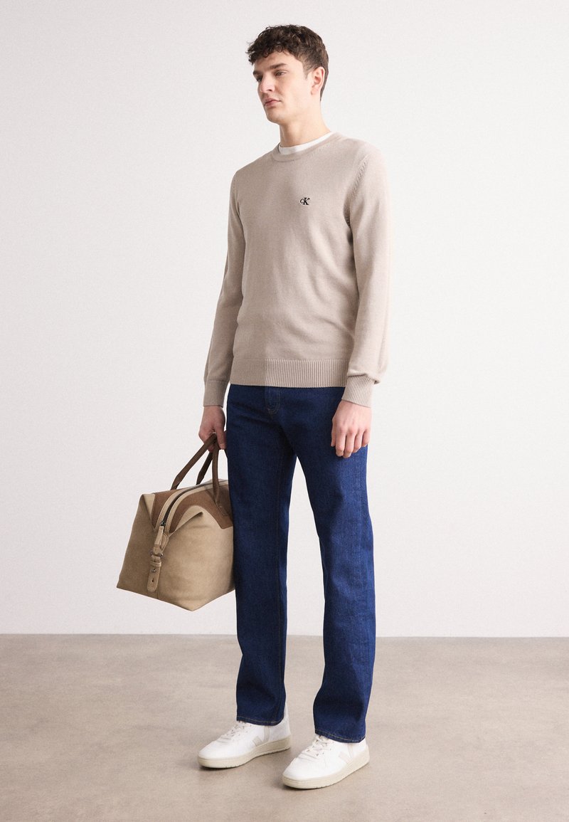 Beżowy sweter dzianinowy z prążkowanym dołem, ciemnoniebieskie dżinsy i białe sneakersy. Beżowa torba z brązowymi akcentami, trzymana w prawej ręce.