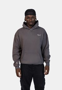 Šedá mikina s klokaní kapsou a vyšitým logem "Crystal". Texturovaná tkanina, volný střih a manžety na rukávech. Nosená s černými cargo kalhotami.