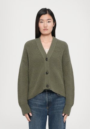 Olive green knit cardigan with a V-neck, three large buttons, and ribbed texture. Loose, cropped fit with long sleeves. Worn over a light top.