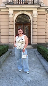 Bela peplum majica s črnimi pikami, svetlo modri raztrgani kavbojke, bele špicate čevlje in majhna bela torbica. Zeleni grmi v ozadju.