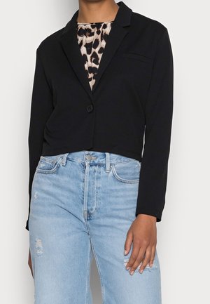 Woman wearing a black cropped blazer over a leopard print top paired with high-waisted light blue ripped jeans against a neutral background.