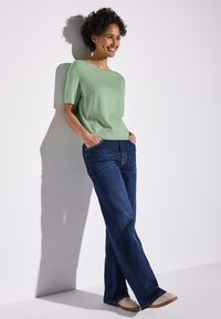 Pull léger vert, à manches courtes, associé à un jean large taille haute bleu foncé. Le modèle se tient avec les mains dans les poches contre un mur blanc.