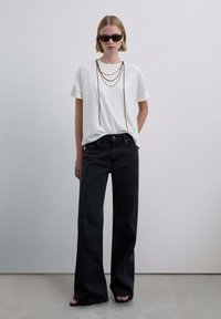 Camiseta blanca, vaqueros negros de talle alto, collar de cadenas en capas y gafas de sol negras, posando sobre un fondo neutro. Sandalias planas visibles.