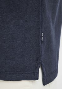 Tums zilā krāsā, izgatavots no struktūrēta auduma, ar sānu nogriežņiem apakšmalā un nelielu logotipa birku kreisajā pusē.