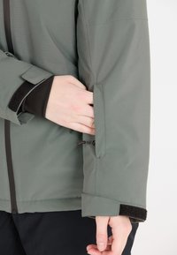 Grüne Jacke mit glatter Textur, ausgestattet mit einer Reißverschlusstasche am Ärmel und schwarzen Klettakzenten am Bündchen.