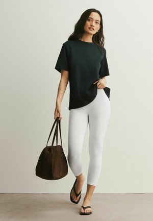 Žena v čiernom tričku s krátkym rukávom, bielych skrátených legínach a čiernych sandáloch, drží veľkú hnedú kabelku v ľavej ruke.