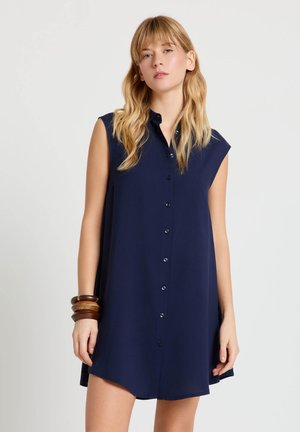 Donna bionda che indossa un vestito blu navy senza maniche con bottoni e braccialetti di legno marroni al polso sinistro, in piedi contro uno sfondo semplice.