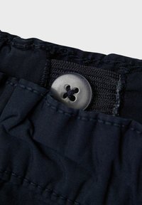 Marineblaue Stoff mit strukturiertem Finish, ausgestattet mit einem runden schwarzen Knopf mit vier Löchern, genähten Nähten und einem gefältelten Abschnitt.