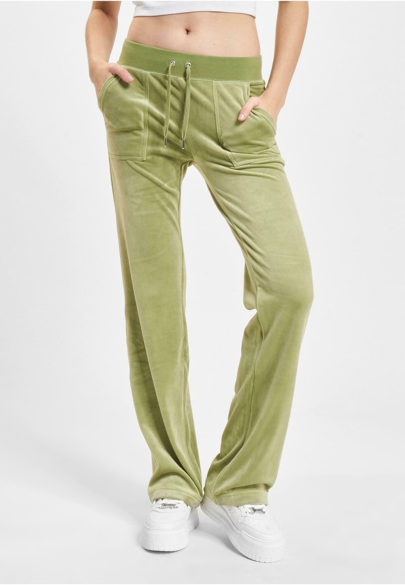 Zielone welurowe dresy z elastycznym paskiem, dwiema przednimi kieszeniami i luźnym krojem. Połączone z białymi trampkami na neutralnym tle.